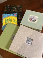 Lot of five hobby and photo albums on wooden surface including a beige fabric photo album, green photo album, floral notebook, lettering workbook, and cross stitch kit box.