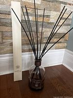 Full view of the large glass diffuser bottle with black reed sticks arranged inside, standing on wooden floor against stone brick wall.