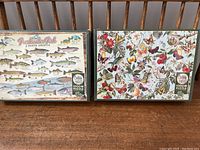Two new in box Cobble Hill puzzles laid on wooden surface. Left puzzle features freshwater fish illustrations; right puzzle features birds, butterflies, and flowers.