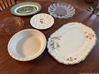 Photo shows six assorted vintage plates and platters of various sizes, materials including ceramic and glass, arranged on a wooden table.