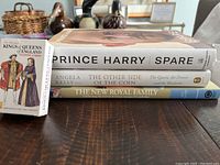 Books stacked horizontally: 'Spare' by Prince Harry on top, 'The Other Side of the Coin' by Angela Kelly in the middle, and 'The New Royal Family' on the bottom, with a deck of playing cards featuring Kings and Queens of England standing upright in front.