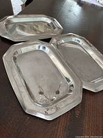 Three rectangular stainless steel trays arranged on a dark wood surface, showing shape, rim style and reflection.