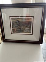 Framed print showing stormy coastal scene with rocks, waves, and windblown tree, against moody sky colors.
