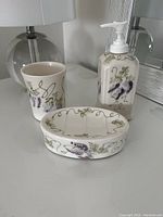 Set of three ceramic bathroom items: soap dispenser with pump, tumbler, and soap dish with floral and vine design.