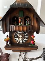 Front view of chalet-style wooden cuckoo clock showing Roman numeral dial, painted figurines on balcony and base, and attached weight chains