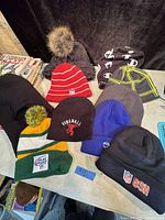 Ten assorted toques and one baseball style cap laid out on a table, showing variety of colors and logos