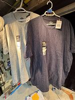 Two men's T-shirts hanging on white plastic hangers, one heather gray and one navy blue, both with tags attached, photographed indoors on a rack.