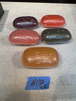 Top-down photo of five wrapped soap bars in various colors: dark purple, red, light pink, dark green, and orange. A blue tape with handwritten "#132 x 5" is visible beneath them.