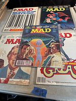 Photo showing five vintage MAD magazines arranged on a surface, highlighting various cover illustrations and the barcode on one cover.