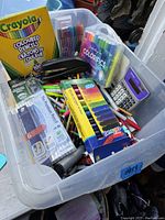 Top-down view of clear plastic bin filled with assorted stationery including colored pencils, pens, markers, and a calculator.