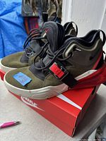 Side view of the olive and black Nike Air Force sneakers with red accents sitting on a Nike shoe box, showing details like the red strap and buckle.