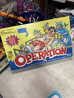 Photo of Operation classic board game box with colorful yellow cover showing cartoon patient on operating table and game details.