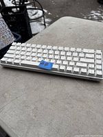 White Royal Kludge mechanical keyboard placed on a concrete surface outdoors, showcasing full keyboard layout and the branding on the bottom right corner.