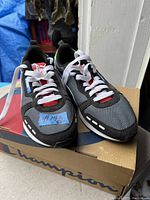 Pair of Puma men's running shoes in black, grey, and white colorway with white laces sitting on a Champion branded shoebox. The shoes appear new and unworn, with blue tags attached to one shoe.