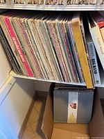 Photo shows a collection of vinyl albums aligned on a shelf, with some stacked in a cardboard box below. Visible titles include 'The Greatest Rock and Roll' and 'Clean Miller Second Trio'.