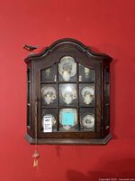 Front view of dark wood wall-mounted display cabinet with the china set visible through glass doors; decorative curved top with a red bird figure on top