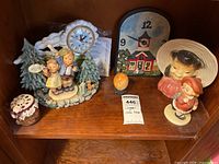 Photo showing Hummel schoolhouse clock figurine with two children and trees, small jar with fabric lid, orange sphere, schoolhouse wall clock, and two ceramic figurines.