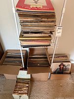 Large stack of vinyl records on metal shelf and boxes on floor, various titles and artists, shows wear, untested condition