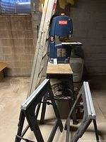Ryobi band saw set on a wooden table with 6ft aluminum ladder leaning against the wall and two black plastic saw horses folded and placed in front