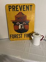 Yellow metal Smokey Bear sign with text 'Prevent Forest Fires' and the bear wearing a ranger hat with 'Smokey' written on it next to a white enamel teapot with red handle and lid knob.