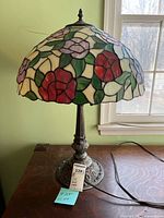 Full view of Tiffany style lamp with floral stained glass shade and ornate metal base on wooden surface near window.