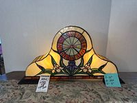 Front view of Tiffany style stained glass lighted clock placed on table showing floral design and clock face.
