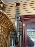 Taller silver-tone metal wind chime with wooden end pieces hanging vertically on a wall beside a red cabinet and wicker furniture.