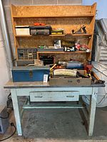 Front view of metal workbench with wooden shelving and assorted tools and toolboxes on top showing drawer and steel top.