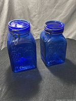 Two cobalt blue glass canisters side by side on a dark surface with light gray fabric background.