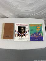 Three books laid flat on a table: left is a brown hardcover book signed by Arnold Schwarzenegger; middle is a white souvenir program with Arnold's photo and bodybuilders from the 1989 Arnold Schwarzenegger Classic; right is a colorful 1990 Arnold Schwarzenegger Classic and Ms. International program featuring Arnold's stylized portrait and bodybuilders.