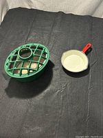 Photo showing cast iron steamer with vented green lid and cast iron fry pan with red handle on black fabric background.