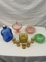 Lot of colored glassware arranged on white cloth background including blue textured bottle with cork, green covered dish, two pink covered bowls, six small amber glasses, and square amber dish.