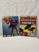 Two DC Comics metal signs shown side by side on white background. Left sign shows Superman with text 'The Man of Steel'. Right sign is vintage Detective Comics cover featuring Batman in action.