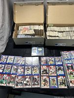 Two cardboard shoe boxes filled with assorted collectible cards, mostly loose and without protective sleeves, with one single card visible on top and displayed football cards in plastic sleeves below the boxes.