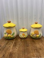 Front view of three ceramic mushroom canisters with yellow lids and mushroom-shaped knobs, shown on wooden surface.