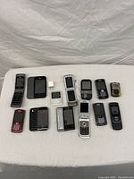 Lot displayed on white fabric surface showing front view of 17 old cell phones and one small adapter piece, various brands and models, mostly candy bar and flip styles
