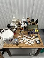 Lot of painting and art supplies arranged on wooden table including gesso container, palette knives, paintbrushes, various bottles and mediums, and a decorative metal tin with sponges.