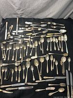 Full spread overview of the lot on a black cloth showing many silver plated cutlery pieces including forks, knives, and spoons with visible wear, patina, and varying styles