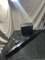 Photo showing black sound bar, wireless subwoofer, power adapter, and cables on black fabric background.