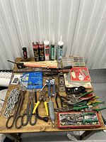 Top-down photo of wooden table showing assorted tools, saw blades in original packaging, caulking tubes, screws, and chains laid out.
