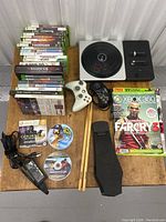 Overview photo showing stacked Xbox and other game cases, accessories including controllers, drumsticks, drum pedal, and power adapter on wooden surface.