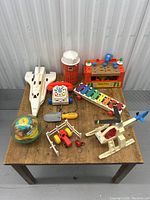 Overview of all vintage Fisher-Price toys on a wooden table showing airplane, tractor with figure, barn silo, telephone, xylophone, helicopter with broken rotor, and small accessories.