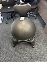 Black inflatable ball chair with backrest and four-legged base on carpeted floor, surrounded by office chairs.