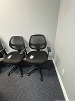 Two black mesh office chairs shown side by side on carpeted floor against gray wall. Seats are cushioned with armrests and five wheeled base.