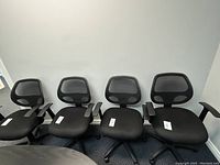 Five black office chairs with mesh backs and padded seats arranged in a row against a wall, showing overall condition and style.