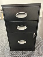 Front view of black three drawer filing cabinet with textured surface and silver oval handles, key inserted in lock on bottom drawer.