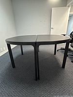 Two round half tables pushed together forming an oval/round conference table in a carpeted office space with a chair, white walls, and closed door.