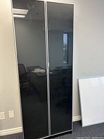 Front view of tall black and grey bookcase with closed glass doors reflecting office surroundings.