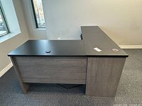 Left side view showing L shape with wood grain base and black top. Visible cable management grommet and power outlet on surface.