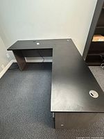 Right hand facing L-shaped executive desk with dark laminated wooden finish on carpeted floor next to white wall.